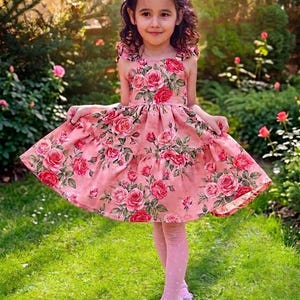 May include: A sleeveless dress featuring a pink base with a vibrant red and pink rose floral pattern, a fitted bodice, and a flared skirt with a tiered hemline. The dress is paired with light pink patterned tights and light pink ballet flats.