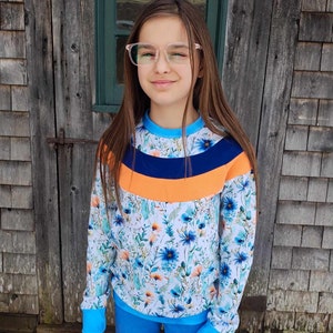 May include: A girl wearing a white, blue, and orange long-sleeved shirt with a floral pattern. The shirt has a blue, orange, and navy blue stripe across the chest. She is wearing blue jeans and clear glasses.