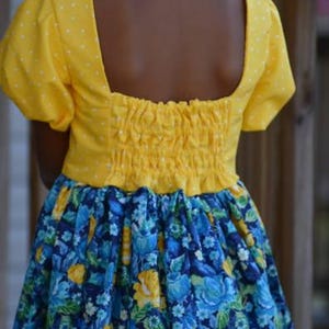 Puede incluir: Un vestido amarillo con escote cuadrado y mangas cortas abullonadas. El corpiño tiene la espalda fruncida. La falda es de estampado floral azul con detalles amarillos y blancos. El vestido está diseñado para una niña.