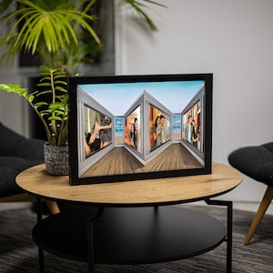 May include: A black framed photo collage featuring multiple images of people. The photos are arranged to create a perspective effect, with a wooden floor and a blue sky visible in the background. The frame is displayed on a round wooden table.