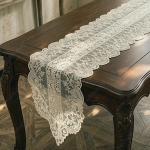 May include: A long, rectangular table runner made of cream-colored lace fabric. The runner features an intricate floral pattern throughout the center and along the edges, which are finished with a scalloped border design that hangs over the table.