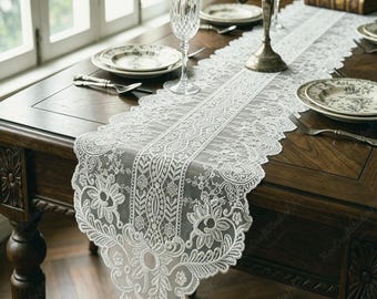 May include: A white lace table runner featuring an intricate floral and geometric pattern with scalloped edges. The runner is laid across a dark wooden table set with white ceramic plates, silver forks, a crystal glass, and a metal candelabra.