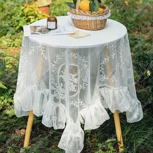 May include: A round white table with a delicate white lace tablecloth. The tablecloth features floral embroidery and a ruffled edge. A small wicker basket with oranges sits on top, along with a jar and a magazine. The table has wooden legs.