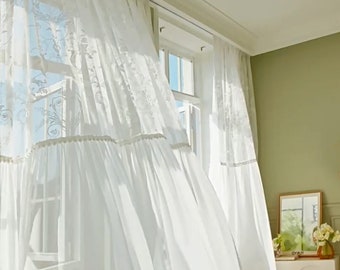 May include: White sheer curtains with delicate lace embroidery hang in a window, blowing gently in the breeze. The curtains have a ruffled bottom edge and are gathered at the top with a white ribbon.
