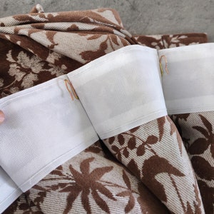 Puede incluir: Primer plano de una tela marrón y blanca con un patrón floral. La tela tiene una cinta blanca cosida a lo largo del borde superior con pequeños ganchos metálicos unidos.