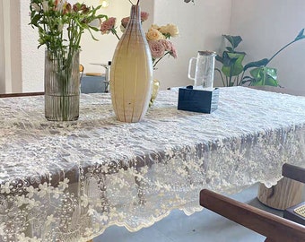 May include: A white lace tablecloth with a scalloped edge, featuring a delicate floral embroidery pattern. The tablecloth is draped over a wooden table with two chairs. A vase with flowers and a glass pitcher are on the table.