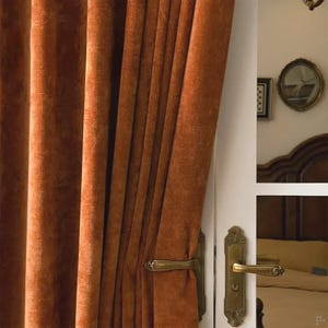 May include: A close-up shot of a deep rust-coloured velvet curtain, with pleats and folds. A brass door handle is visible, suggesting a classic interior. The curtain is partially drawn, offering a peek into a room, creating a sense of depth and luxury.