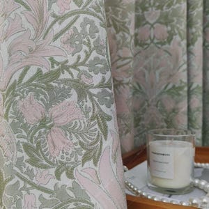 Könnte beinhalten: Nahaufnahme eines Stoffes mit floralem Muster in Rosa, Grün und Weiß. Das Design zeigt stilisierte Blumen und Blätter. Eine brennende Kerze in einem Glas steht auf einem Holztablett mit einer Perlenkette.