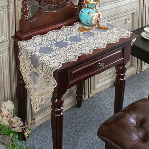 May include: A delicate, off-white lace table runner with a gold embroidered floral pattern. The runner is draped over a dark wood table with a drawer and turned legs.