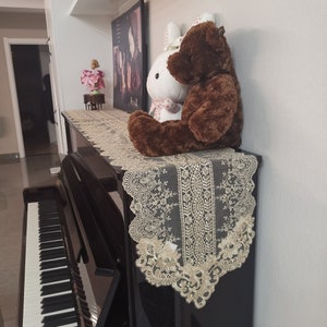 Puede incluir: Un piano vertical negro con un camino de mesa decorativo de encaje beige. Un oso de peluche marrón y un conejito blanco con una bufanda floral rosa están encima. También se ven una imagen enmarcada y un pequeño objeto decorativo.