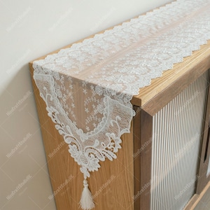 May include: A white lace table runner with a floral design and a tassel. The runner is draped over a wooden cabinet. The lace has a delicate, intricate pattern, adding a touch of elegance to the decor.