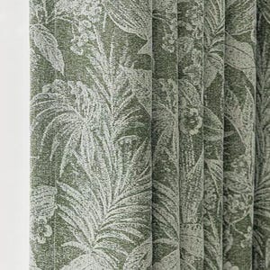 Pode incluir: Close-up de uma cortina estampada em verde e branco. O tecido apresenta um design repetido de grandes folhas tropicais e elementos florais. A cortina é dobrada em pregas verticais, mostrando a textura e profundidade do material.