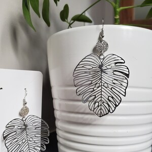 Puede incluir: Un par de pendientes de plata con un delicado dise&ntilde;o de hoja recortada y un peque&ntilde;o abalorio circular de plata brillante. Los pendientes se muestran sobre una superficie blanca.