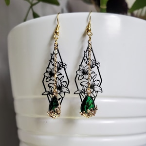 May include: A pair of dangle earrings with a black floral design and a green teardrop-shaped gemstone. The earrings are made of metal and have a gold finish.