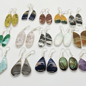 May include: A selection of gemstone earrings with silver hooks. The earrings showcase a variety of natural stones in different shapes and colours, including green, blue, brown, and purple. The earrings are displayed on a white surface.