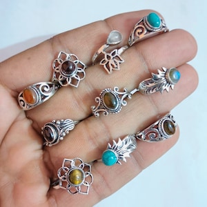 Può includere: Un set di nove anelli d'argento con diverse intarsi di pietre preziose. Gli anelli presentano diversi design, tra cui motivi floreali, solari e a foglia. Le pietre preziose includono occhio di tigre, turchese e pietra di luna.