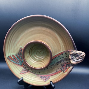 Könnte beinhalten: Eine Keramik-Servierschale mit Fischdesign. Die Schale ist hauptsächlich beige und grün, mit einem braunen Rand und einer kleinen, kreisförmigen Dip-Schale in der Mitte. Der Fisch ist mit roten, grünen und schwarzen Akzenten dargestellt.