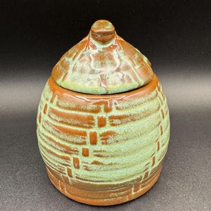 May include: A handmade ceramic jar with a lid. The jar is a light green color with brown horizontal and vertical lines. The lid is brown with a small knob on top. The jar is a unique piece of pottery.