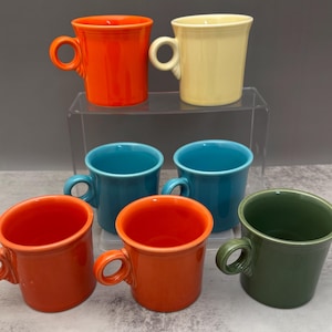 May include: Six ceramic mugs in a range of colours: orange, yellow, teal, and green. Each mug has a rounded handle and a slightly flared rim. The mugs are arranged on a clear, tiered surface.