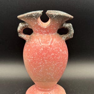 Könnte beinhalten: Eine Keramikvase mit einem korallenrosa Körper und einer weiß gesprenkelten Glasur. Das Gefäß hat zwei seitliche Griffe und einen ausgestellten, gewellten Rand mit einem dunkelgrau-weißen Sprenkelmuster, das sich bis zu den Griffen erstreckt.