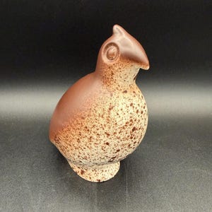 Może przedstawiać: Ceramiczna figurka ptaka z gładką, jednolicie brązową górną częścią oraz nakrapianym, kremowym tułowiem z brązowymi plamkami. Ptak posiada mały, spiczasty dziób, okrągły detal oka oraz zaokrąglony, pękaty kształt osadzony na stabilnej podstawie.
