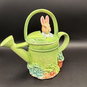 May include: A light green ceramic watering can featuring a sculpted rabbit figure emerging from the lid. The base is decorated with relief carvings of leafy green vegetables and small orange carrots, complemented by a curved handle and spout.