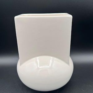 Puede incluir: Un jarrón de cerámica de color blanco roto con un diseño estructural único. La base tiene una forma redondeada y bulbosa que se transforma hacia arriba en una sección superior rectangular y plana con una boca ancha, creando una silueta geométrica distintiva.