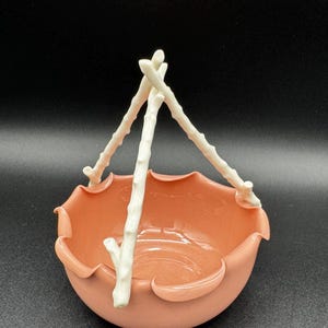 May include: A peach-colored ceramic bowl with a scalloped rim and three white branch-like handles. The bowl has a smooth, glossy finish and is set against a dark background. The handles are arranged to form a hanging basket design.