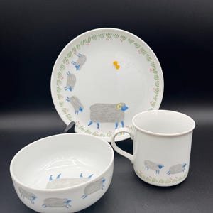 May include: A white ceramic dish set featuring a plate, bowl, and mug. Each piece is decorated with a whimsical design of gray sheep with blue accents and a green border. The set is ideal for serving meals.