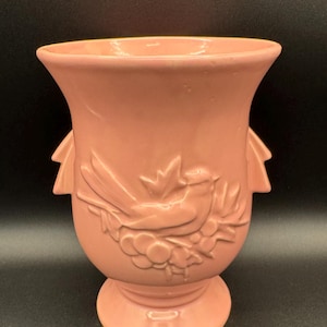 Può includere: Un vaso vintage in ceramica rosa con un motivo a rilievo di uccello e fogliame. Il vaso ha una parte superiore svasata, una base a piedistallo e due manici laterali. L'uccello è appollaiato su un ramo con foglie e bacche.