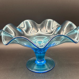 May include: A translucent light blue glass compote dish featuring a wide, ruffled, and scalloped rim. The bowl is supported by a short, sturdy pedestal base that flares out into a circular foot, creating a smooth, elegant, and curved silhouette.