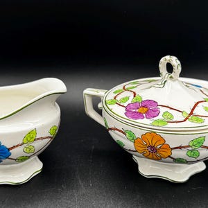 May include: Cream-colored ceramic creamer and covered sugar bowl set. The set features a floral design with blue, orange, and purple flowers, green leaves, and brown vines. The rims and handles are trimmed in green.