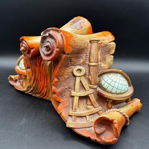 Puede incluir: Un sujetalibros detallado de estilo vintage con un diseño de pergamino enrollado, un mapa, un globo terráqueo e instrumentos de navegación. El sujetalibros es principalmente naranja y marrón con detalles en beige y azul, probablemente de cerámica o resina.