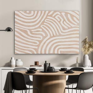 Può includere: Una grande opera d'arte rettangolare su tela con sfondo beige e design a linee bianche curve. L'opera è appesa sopra un tavolo da pranzo rotondo in legno con piatti e sedie neri. Sono visibili anche vasi decorativi e una lampada.