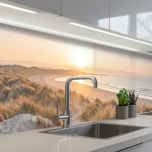 May include: A contemporary kitchen sink with a chrome tap, set against a scenic backdrop of a beach at sunset. The image features a stainless steel sink, a tap, and small potted plants. The background shows a beach with dunes and the ocean.