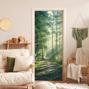 Op de afbeelding: Een deur met een bosscène. De afbeelding toont een pad dat leidt naar een zonnig bos met hoge bomen. De deur is omlijst door een witte rand en maakt deel uit van een kamer met een bank, kussens en decoratieve items.