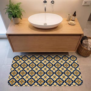 Op de afbeelding: Een rechthoekige badmat met een herhalend patroon van marineblauwe en gele bloemmotieven op een crèmekleurige achtergrond. De mat ligt voor een houten wastafel met een witte ovale wastafel. Een varen in een pot staat links.
