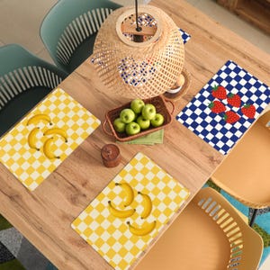 May include: Overhead shot of a wooden table set for a meal. Yellow and white checkered placemats with bananas and blue and white checkered placemats with strawberries are visible. A woven basket holds green apples. A woven light fixture hangs above.