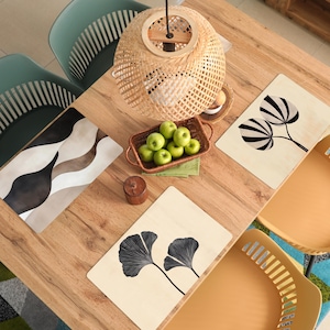 May include: Overhead view of a wooden dining table with decorative placemats, a woven basket of green apples, and a woven pendant light. The placemats feature abstract designs and botanical illustrations. The table is surrounded by teal and yellow chairs.