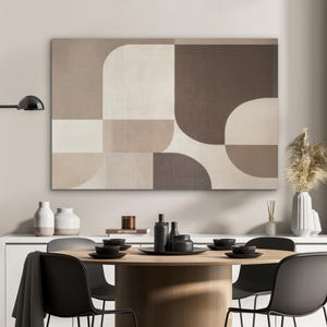 Może przedstawiać: Abstrakcyjna geometryczna sztuka na płótnie w odcieniach beżu, jasnobrązowego i brązu. Grafika przedstawia nowoczesny design z zakrzywionymi i prostokątnymi kształtami. Płótno jest wystawione nad stołem jadalnym z czarnymi naczyniami i krzesłami, tworząc współczesną jadalnię.