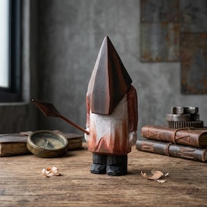 May include: A hand-carved wooden figure of a person wearing a dark brown pyramid-shaped head covering and a red and white tunic, holding a spear. The figure stands on a wooden surface. A compass and books are in the background.