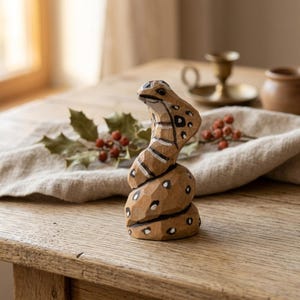 May include: A hand-carved wooden snake figurine with a detailed design. The snake is painted with brown, white, and black accents. The figurine is posed in a coiled position, with its head raised. The snake is on a wooden table.