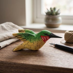 May include: A hand-carved wooden hummingbird sculpture. The bird features green, yellow, and red painted details, with a black beak. The wings are outstretched, and the sculpture is set on a wooden surface.