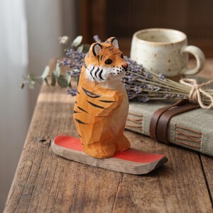 Könnte beinhalten: Handgeschnitzte Holz-Tigerfigur mit orangefarbenen und schwarzen Streifen, die auf einer roten Holzbasis steht. Der Tiger wird in einer stehenden Pose dargestellt. Die Figur steht auf einer rustikalen Holzoberfläche.