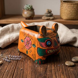 May include: Orange wooden planter in the shape of a tiger with a floral design. The planter has a rectangular shape with a tail and ears. The tiger's face is painted with colorful details, including blue eyes and a red mouth.