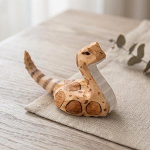 Op de afbeelding: Een houten slangenbeeldje met een lichtbruin lichaam en donkerbruine vlekken. De slang is gesneden met gedetailleerde schubben en een gebogen houding, rustend op een neutraal gekleurd linnen oppervlak. De kop is omhoog en de staart is gekruld.