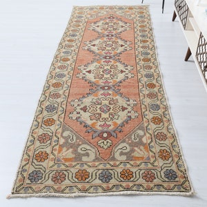 May include: A vintage runner rug with a cream background and a repeating pattern of floral motifs in shades of pink, orange, and blue. The rug has a fringed edge.
