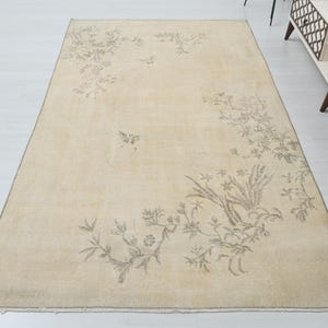 May include: Beige rectangular rug with a floral design in shades of gray. The rug features a central floral pattern with additional floral and bird motifs scattered across the surface. The rug is placed on a white floor.