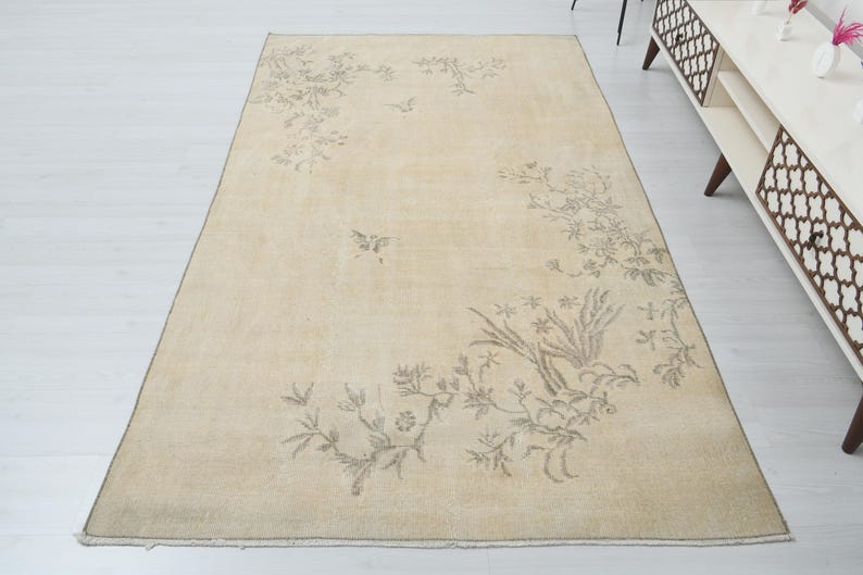 May include: Beige rectangular rug with a floral design in shades of gray. The rug features a central floral pattern with additional floral and bird motifs scattered across the surface. The rug is placed on a white floor.