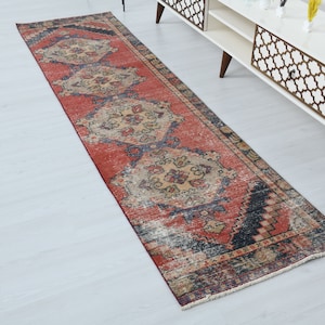 May include: A red, blue, and beige vintage runner rug with a floral pattern. The rug has a fringed edge and is laid out on a white floor.